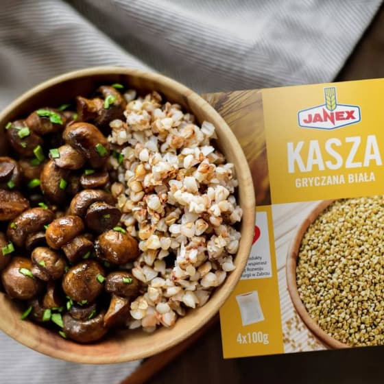 Janex Kasza Gryczana Biała w opakowaniu 4x100g obok drewnianej miski z ugotowaną kaszą gryczaną i podsmażanymi pieczarkami posypanymi szczypiorkiem.