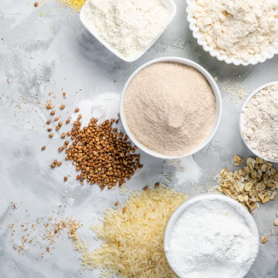Różne rodzaje mąk i ziaren w białych miskach na szarym tle, w tym mąka pszenna, mąka owsiana, ryż i ziarna gryki.