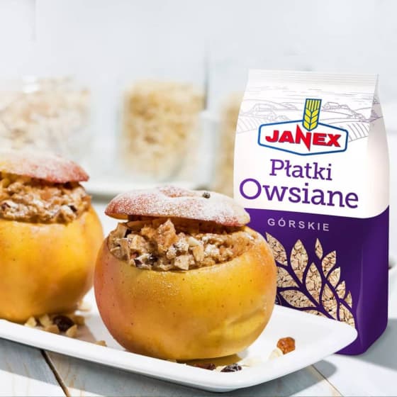 Janex Płatki Owsiane Górskie w białym i fioletowym opakowaniu, obok dwa jabłka nadziewane płatkami owsianymi i bakaliami na białym talerzu.