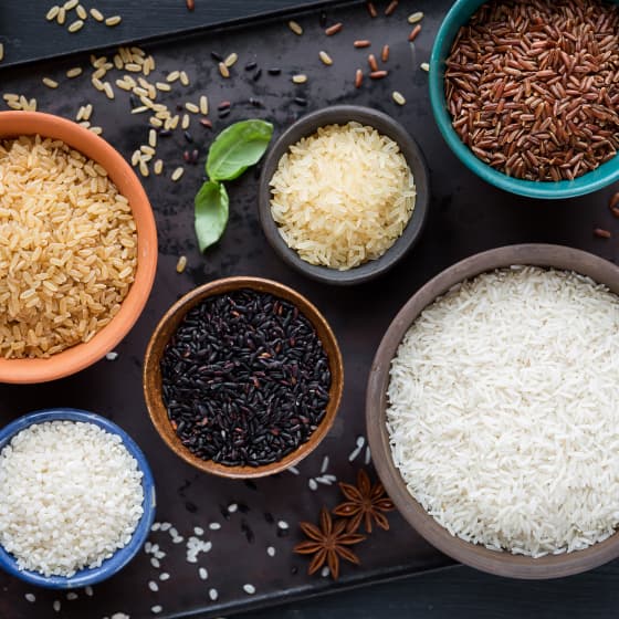 Miska z różnymi rodzajami ryżu: biały, brązowy, czarny, dziki i parboiled, ułożone na ciemnej powierzchni z zielonym liściem i anyżem gwiazdkowatym.