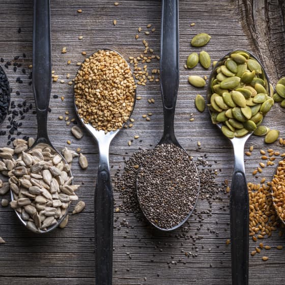 Cztery metalowe łyżki z różnymi nasionami: słonecznik, sezam, chia i pestki dyni na drewnianym tle.